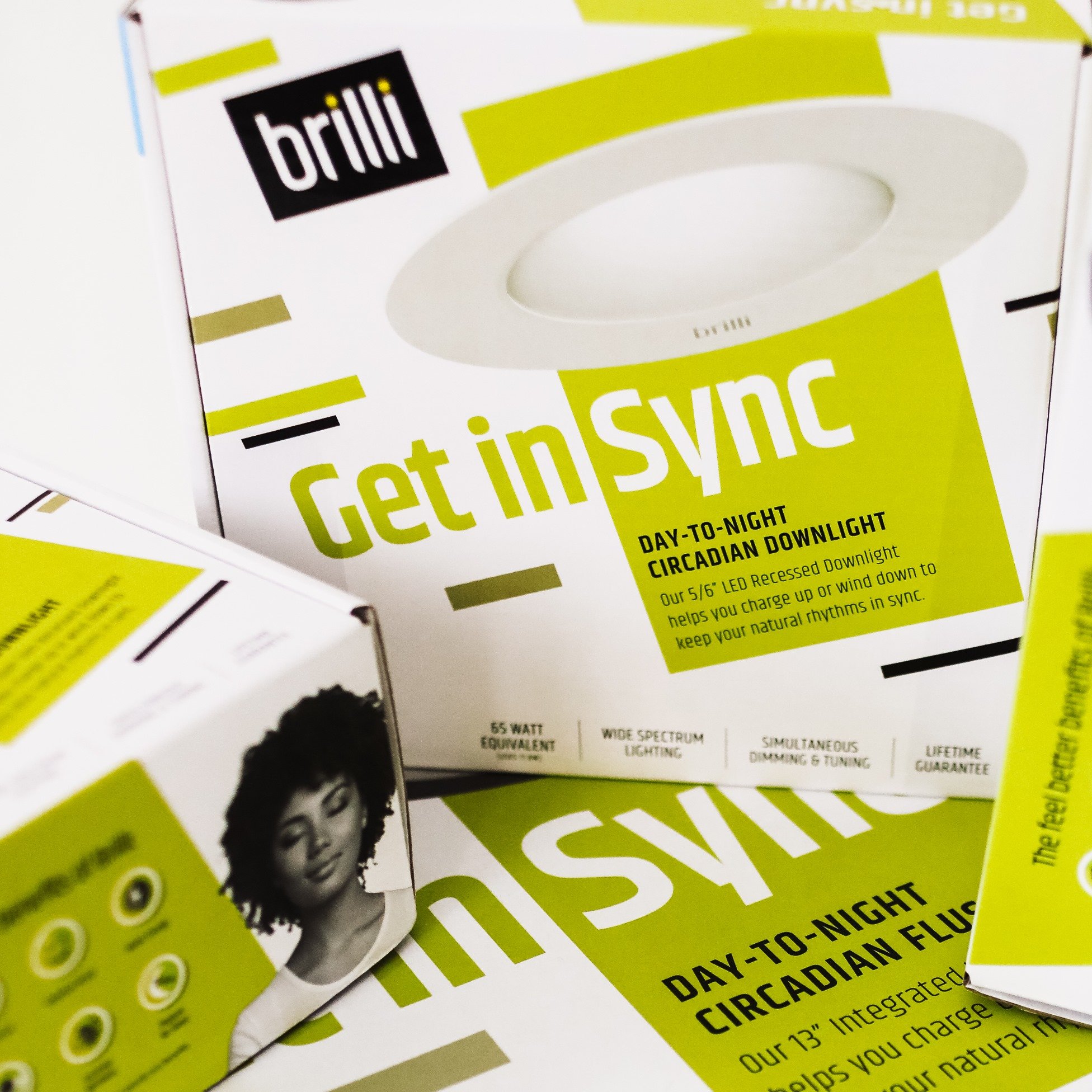 Boxes of Brilli Get in Sync circadian lighting products, including a 5/6 inch recessed daylight and a 13 inch integrated fluorescent light with promotional text.