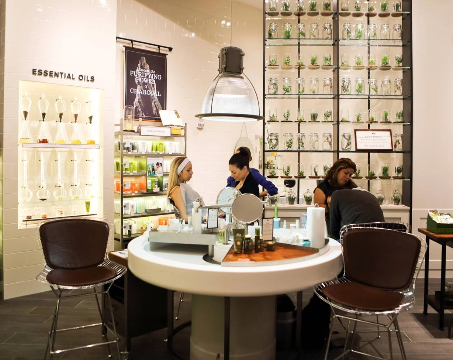 Interior of a retail store with a white round table in foreground and two brown chairs. Behind, customers and staff are behind a counter, with a wall of glass jars filled with green plants on the right. Shelves display essential oils and skincare pro