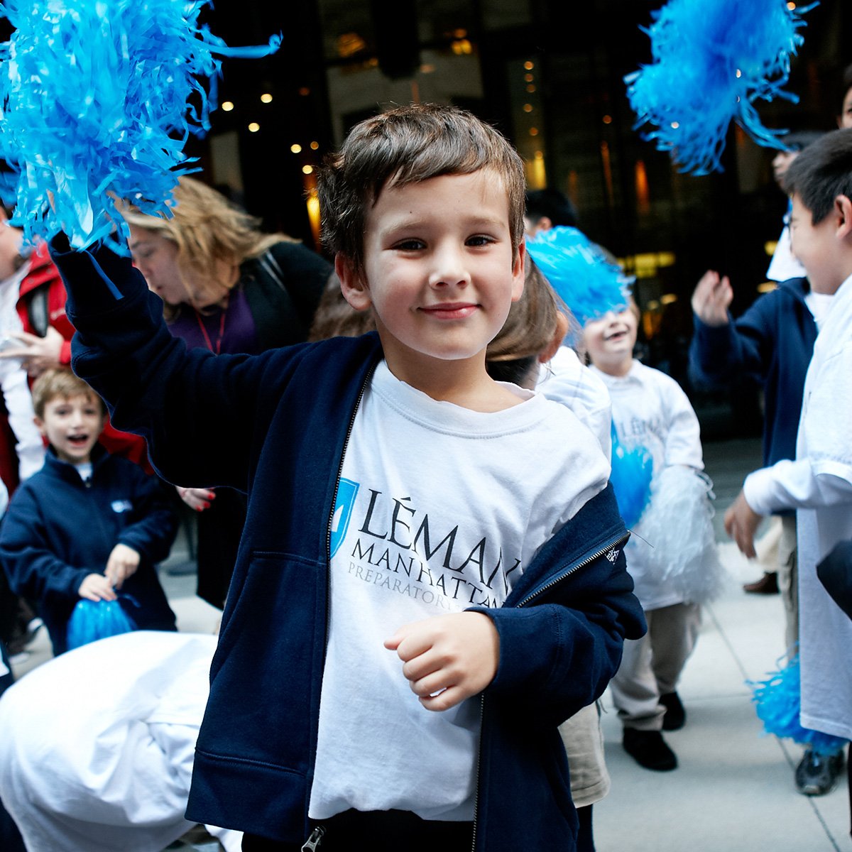 Young boy smiling and waving blue pom-poms at a parade or celebration with other children and adults in the background.