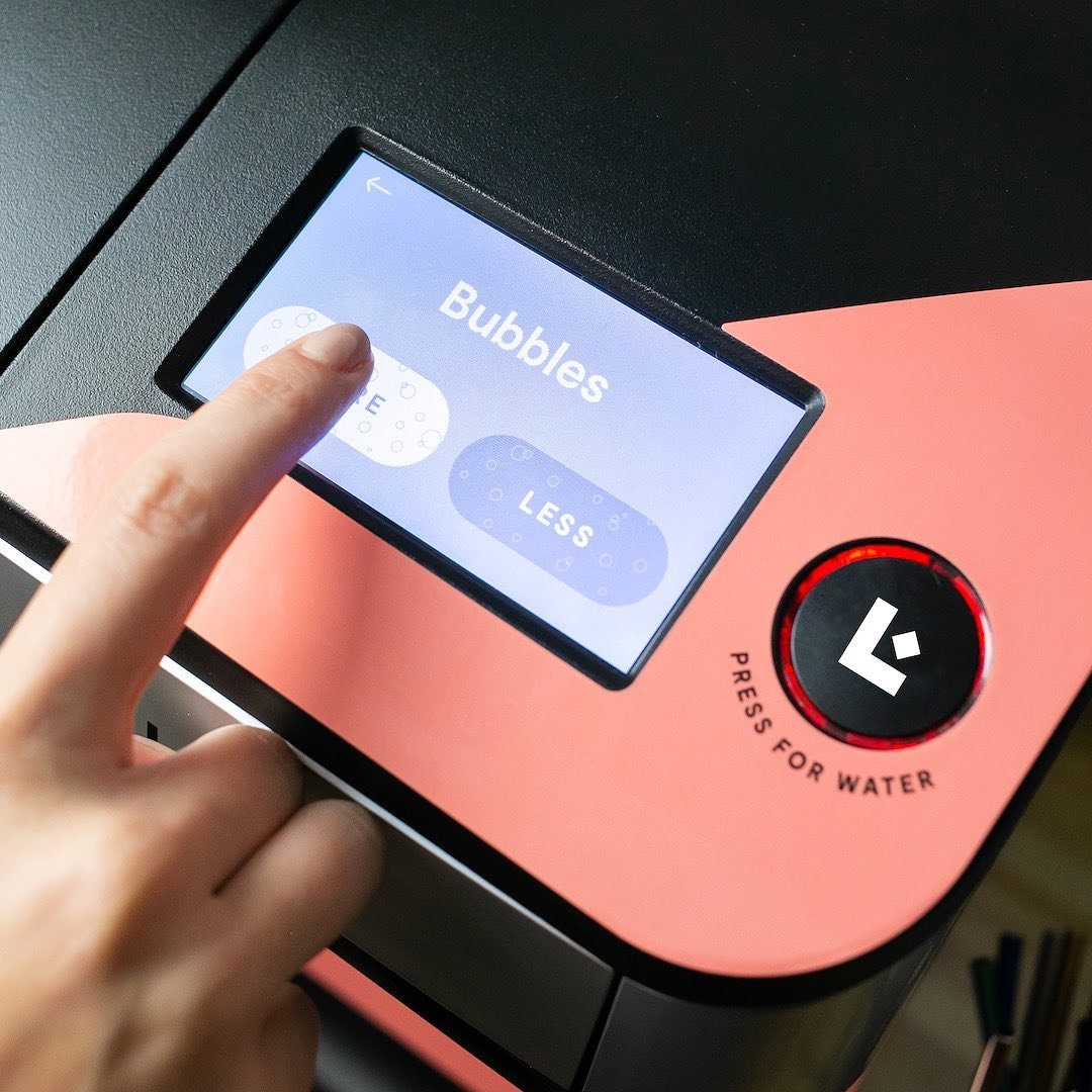 A person pressing a touchscreen button labeled 'Less' on a bubble tea machine with a pink and black exterior. The screen displays options for bubbles.