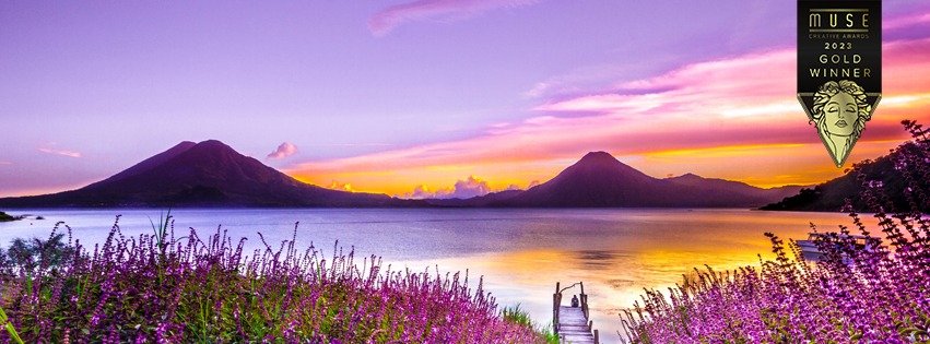 A scenic lake view during sunset with purple and yellow sky, mountains in the background, purple flowers in the foreground, and a small dock leading into the water. A gold award badge in the top right corner indicates a 2023 Muse Creative Awards Gold