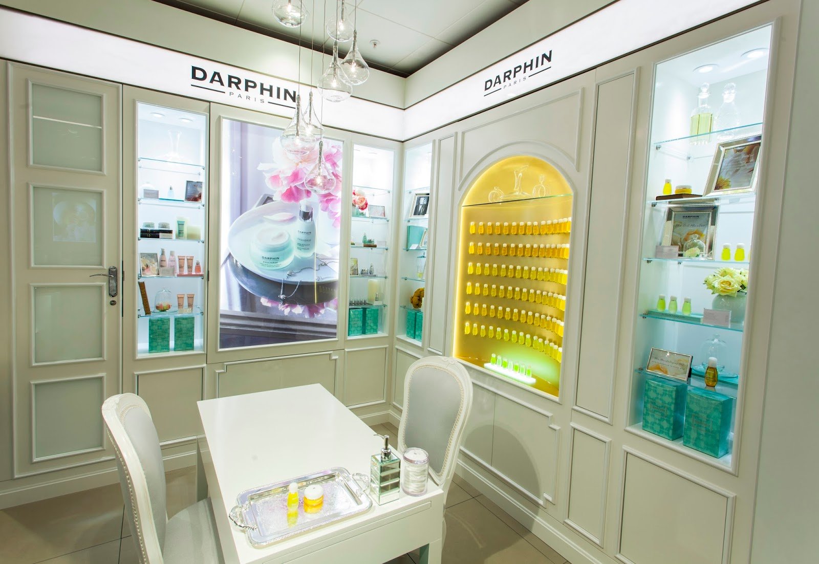 Interior of a cosmetic or skincare shop with display shelves holding bottles of products, a white table with a tray and containers, and an illuminated section with yellow bottles, branded with Darphin.