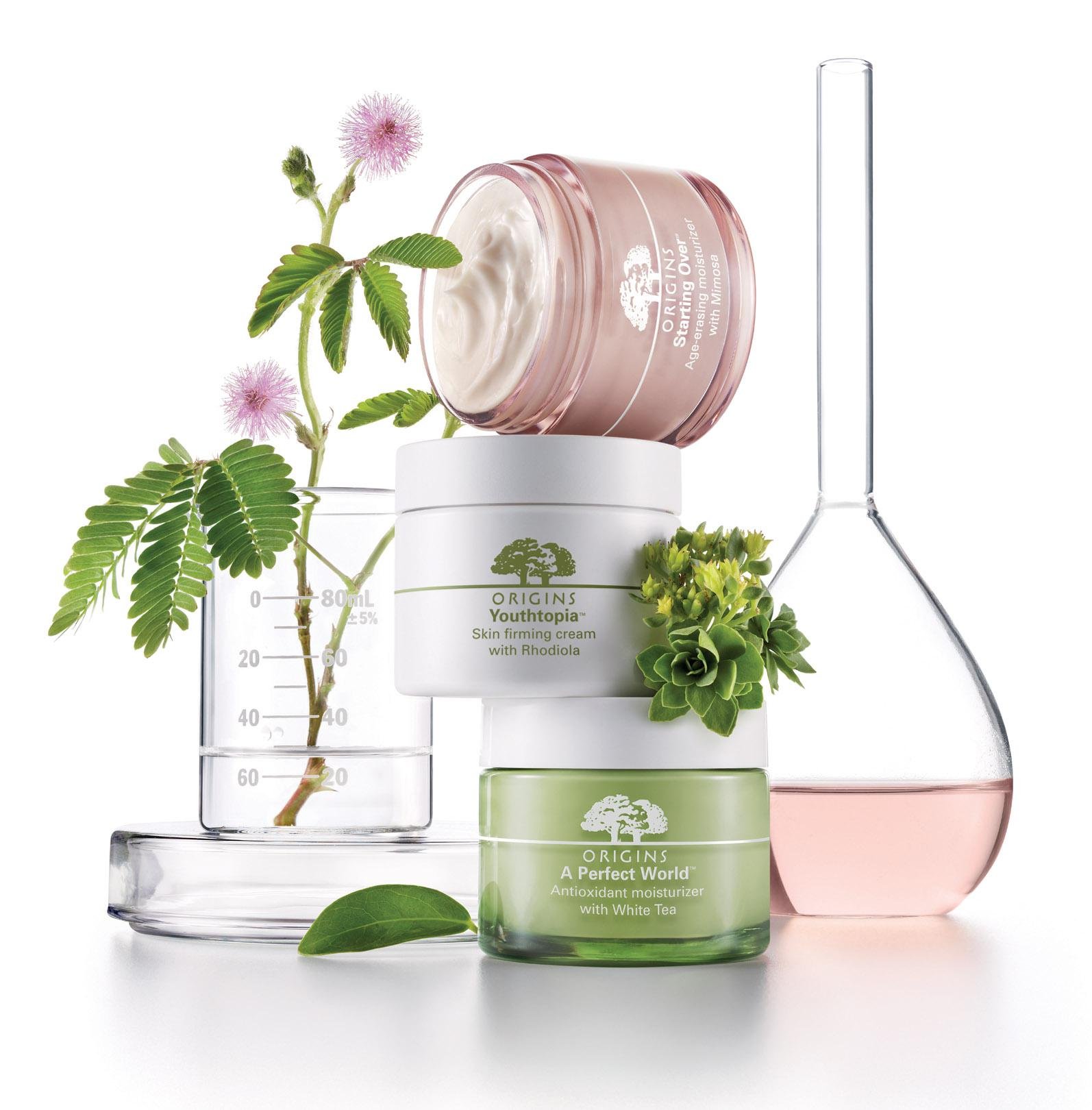 Stacked skincare products including a white jar labeled 'Youthtopia', a pink jar labeled 'Starting Over', and a green jar labeled 'A Perfect World', with green leaves, a small plant, a glass beaker, and a glass flask in the background.