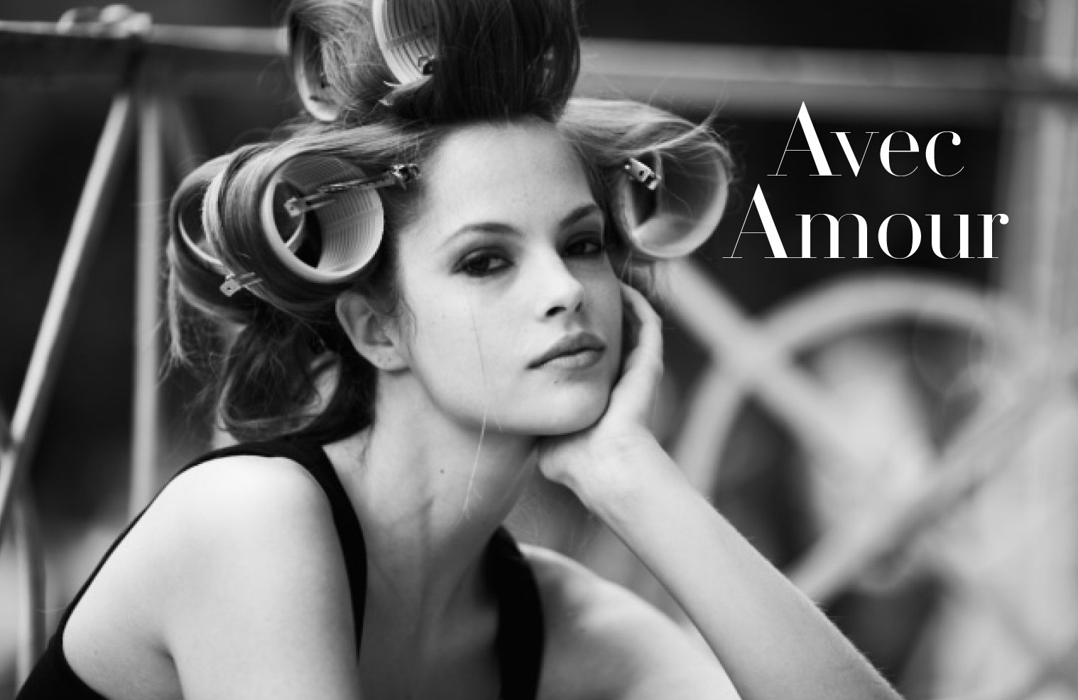 Black and white photo of a woman smiling with large hair rollers in her hair, resting her head on her hand, with the words 'Avec Amour' written on the right side.